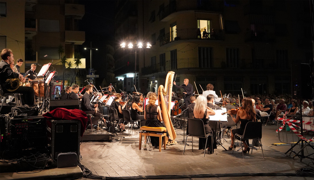 Piazza di un borgo storico, pubblico seduto, orchestra con strumenti a fiato e archi durante esibizione serale estiva, immagine di approfondimento